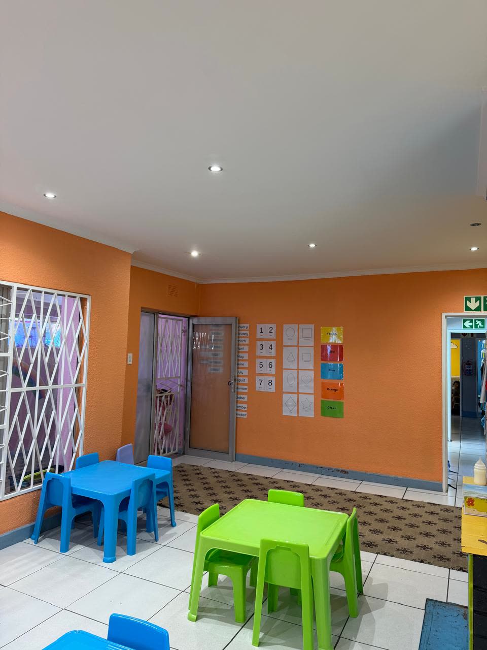 A bright classroom with low tables and chairs, a numbers and shapes wall chart and a marked fire exit